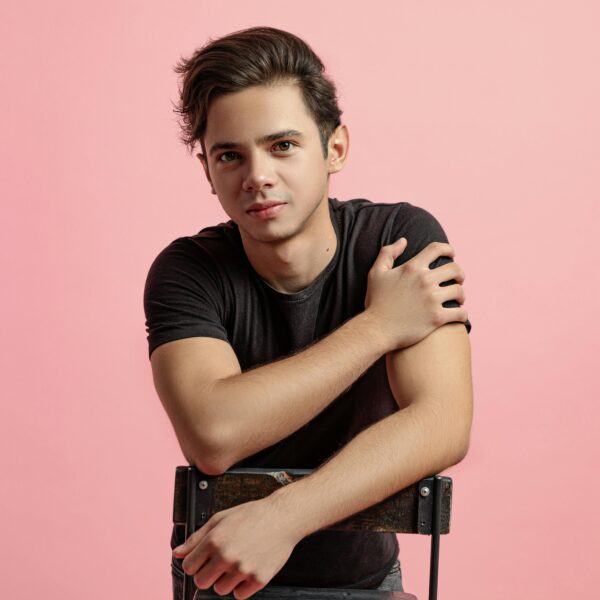 Handsome young sporty male in casual clothing with dark short hair touching shoulder and confidently looking at camera while leaning to chairback on pink background